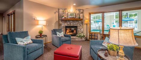 A cozy living room with two blue armchairs, a red ottoman, a stone fireplace, and a large window with a view of a deck.