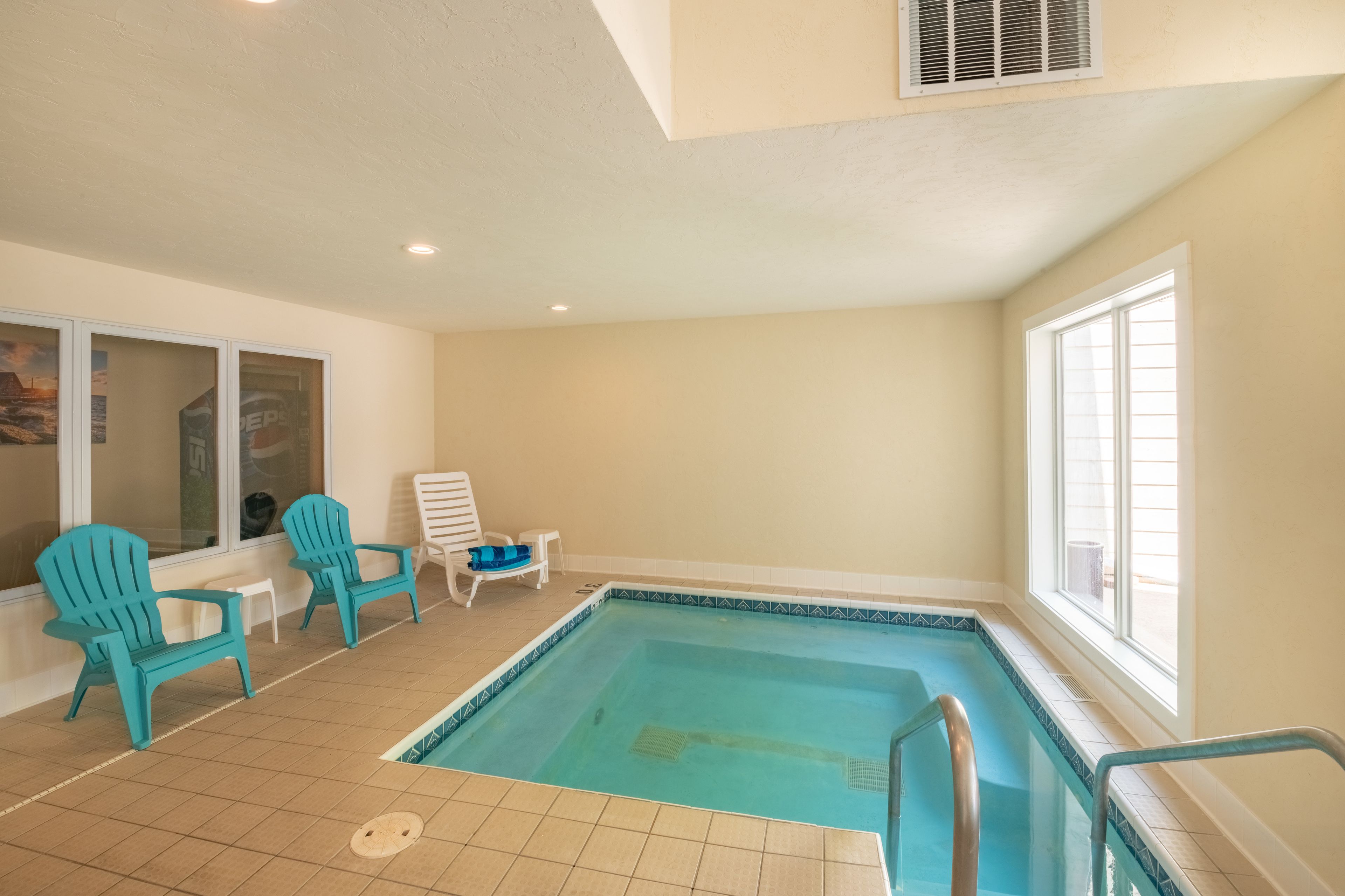 An indoor swimming pool with turquoise chairs and a small table, surrounded by beige walls and large windows.
