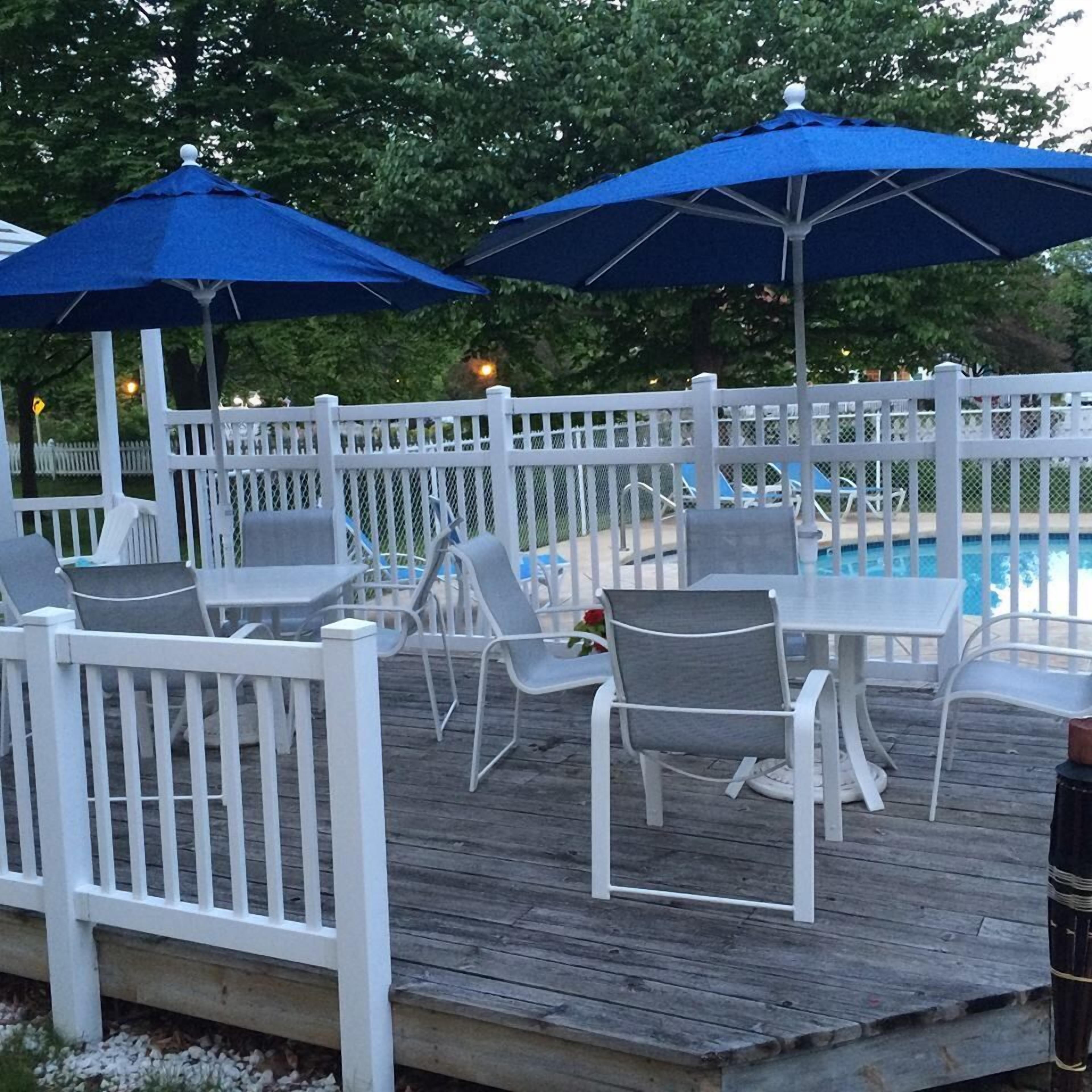 Une terrasse en bois avec des rampes blanches, un espace extérieur aménagé avec des tables et des chaises, et deux parasols bleus.
