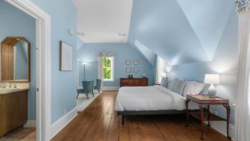 Une chambre avec un plancher en bois, un lit, une table de chevet, une commode, une chaise et une fenêtre avec des rideaux.