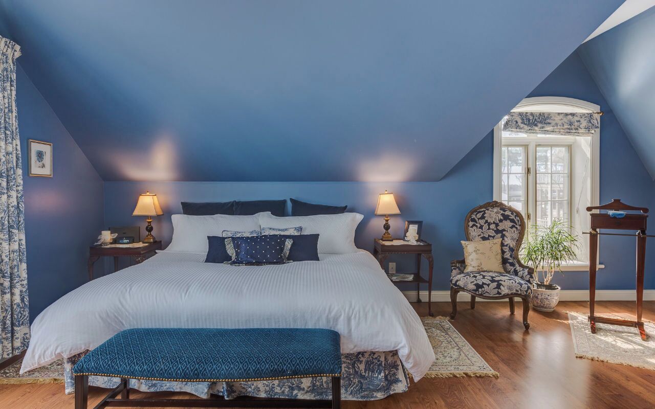 A bedroom with a large bed, two bedside tables with lamps, a chair, a small table, and a window with curtains.