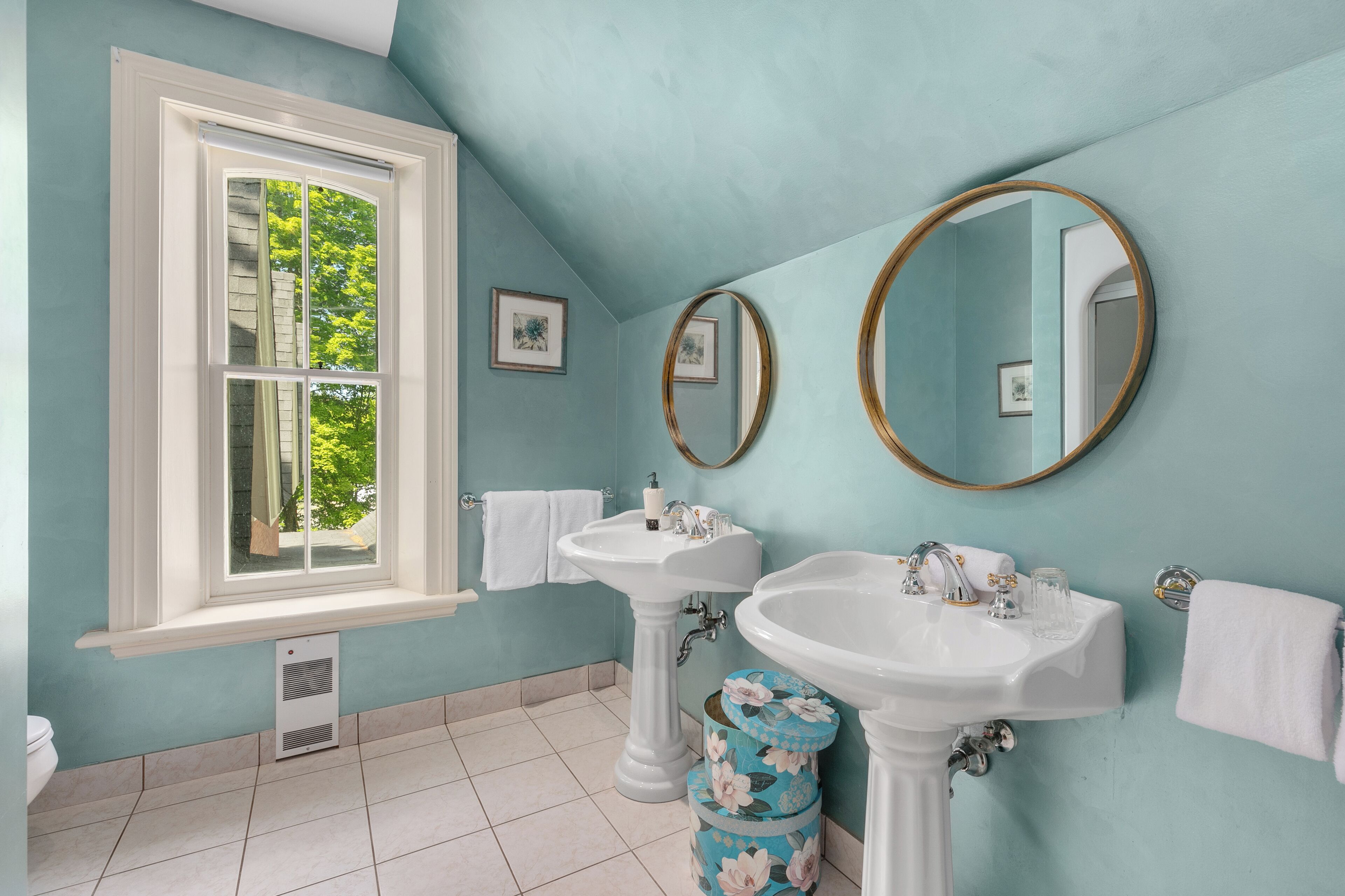 Une salle de bain avec deux lavabos sur pied, une fenêtre donnant sur la verdure et un sol carrelé.