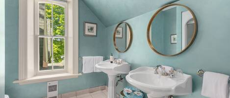 A bathroom with two pedestal sinks, a window with a view of trees, and a tiled floor.