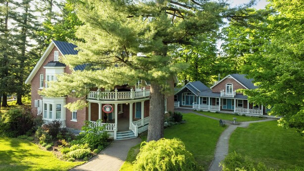 A large, two-story house with a wrap-around porch, surrounded by lush greenery and trees. - Manoir Sweetsburg (Cowansville)