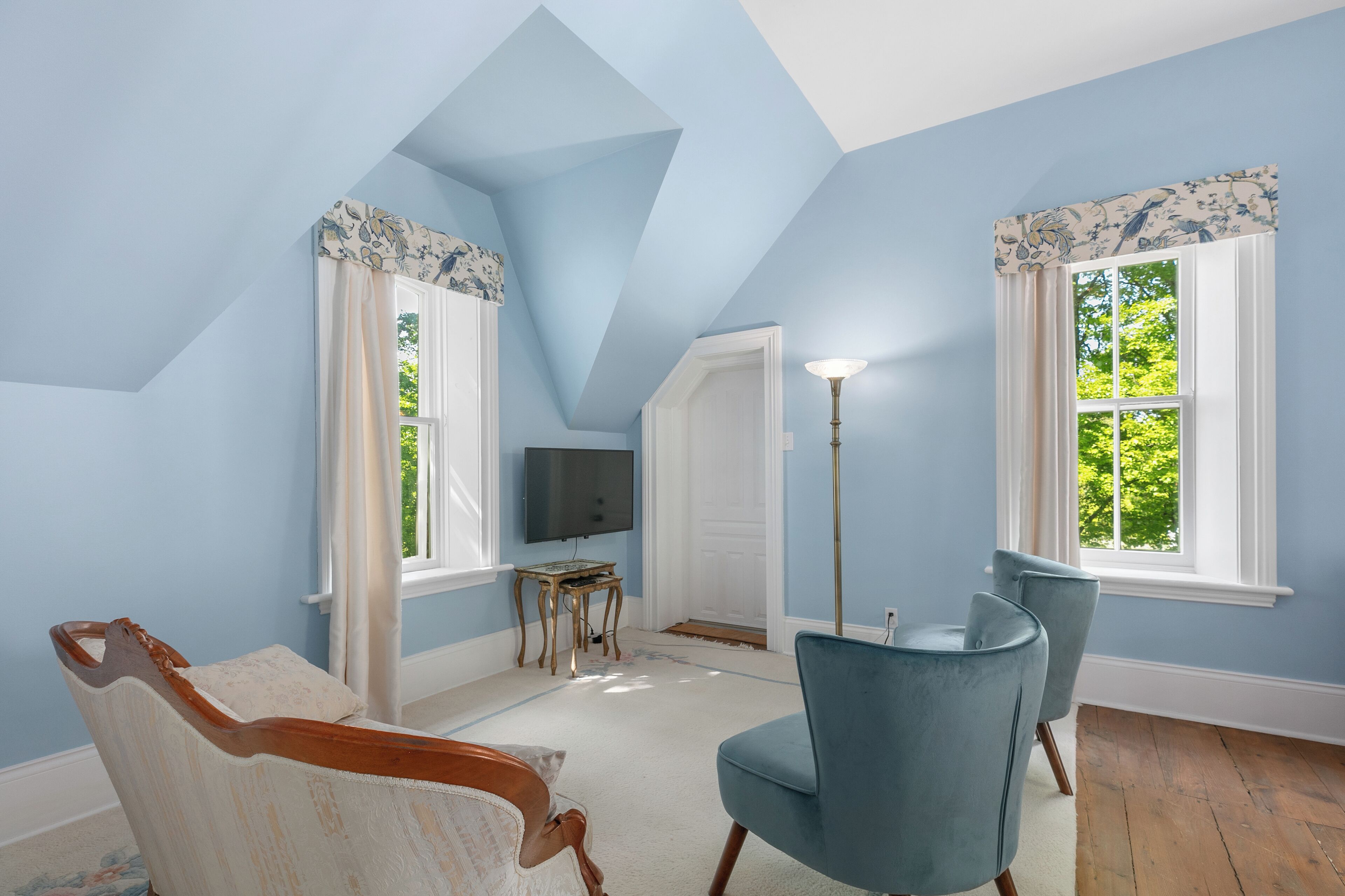 Una habitación con techo inclinado, dos ventanas con cortinas florales, un televisor sobre una mesita y una silla azul.