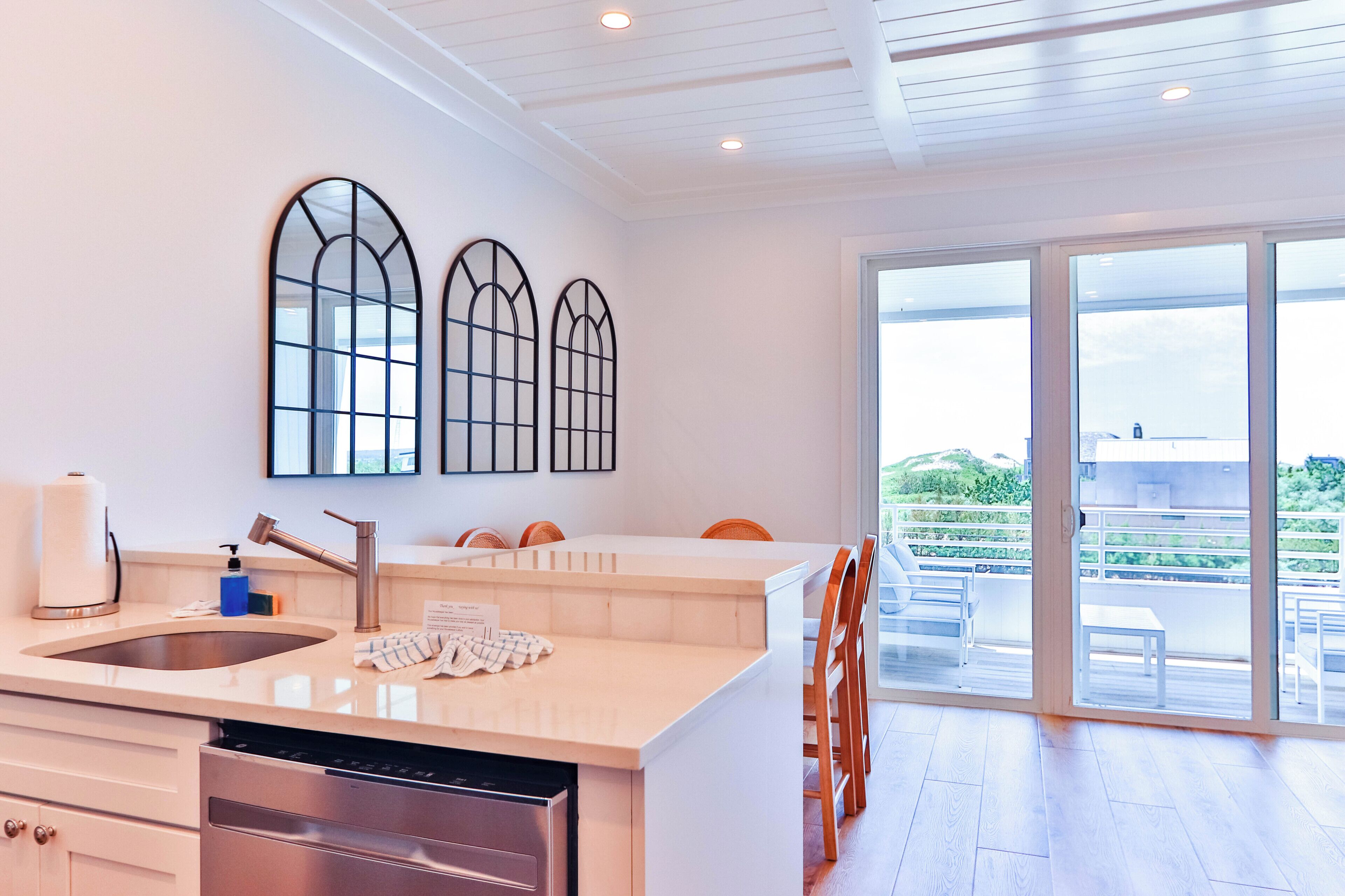 A modern kitchen with a dishwasher, a sink, and a large island with chairs. There are three arched windows on the wall and a sliding glass door leading to an outdoor area.