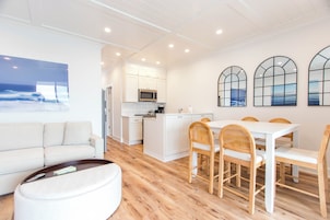 A modern kitchen with white cabinets and a microwave, a dining area with a white table and wooden chairs, and a large window with a view of the ocean.