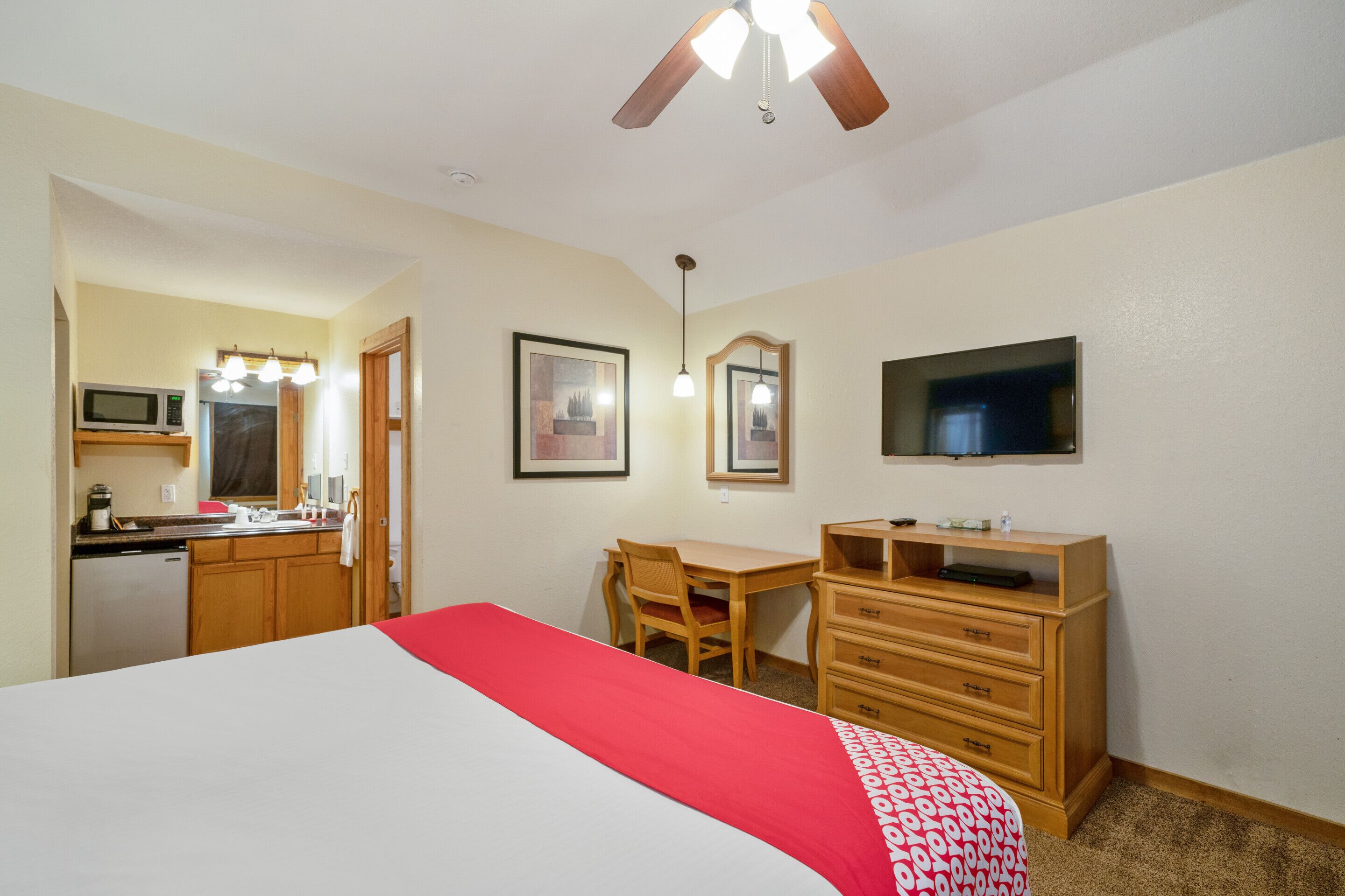 Habitación de hotel con cama, escritorio, silla, cómoda, televisión, ventilador de techo y kitchenette con microondas.