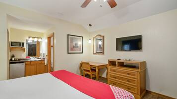 Habitación de hotel con cama, escritorio, silla, cómoda, televisión, ventilador de techo y kitchenette con microondas.