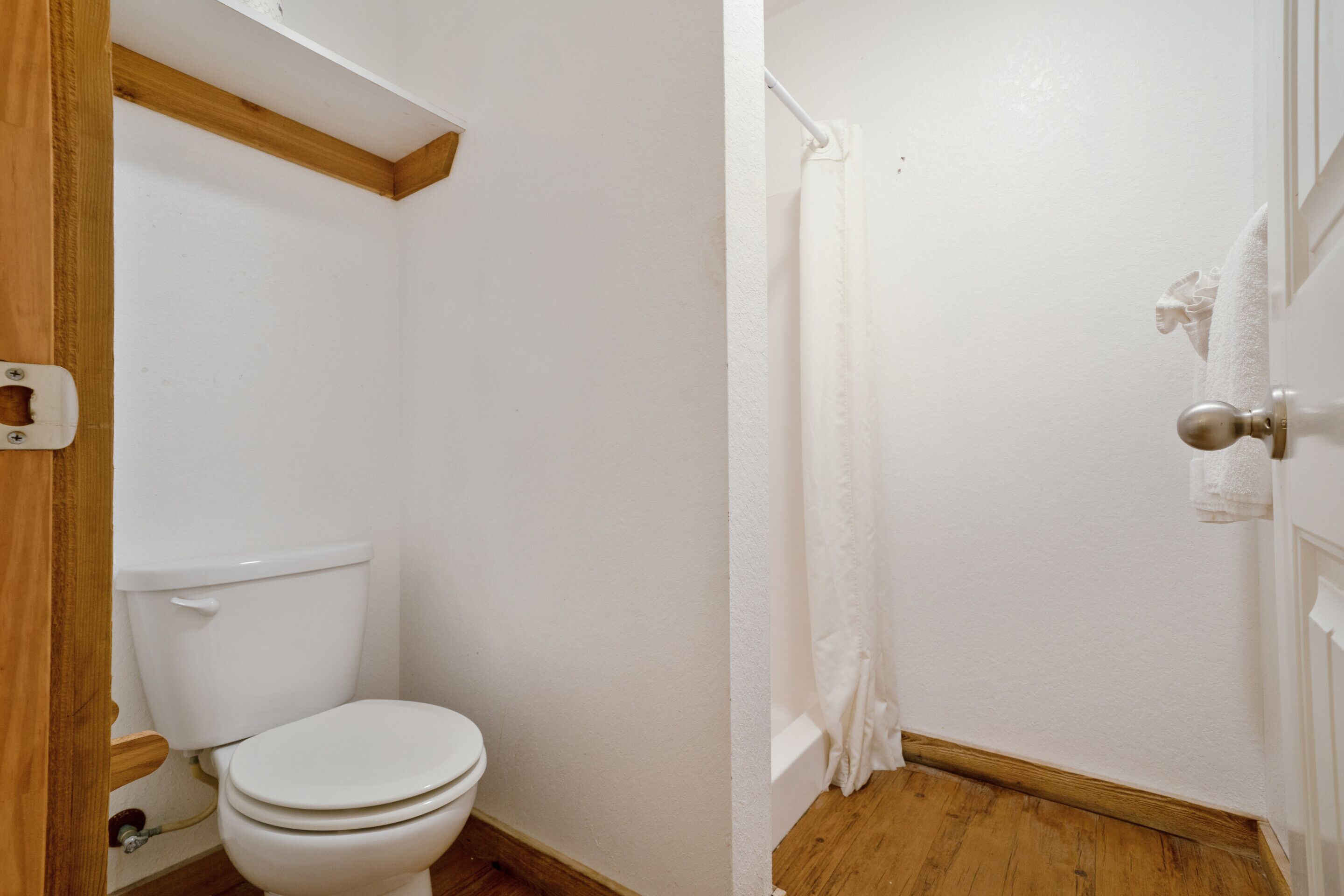 Et badeværelse med toilet, trægulv, hvide vægge og en brusekabine.
