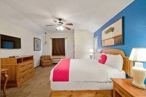 A hotel room with a bed, a desk, a chair, a ceiling fan, a painting, and a lamp.
