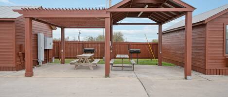 A covered patio area with picnic tables and a barbecue grill, surrounded by wooden fences.