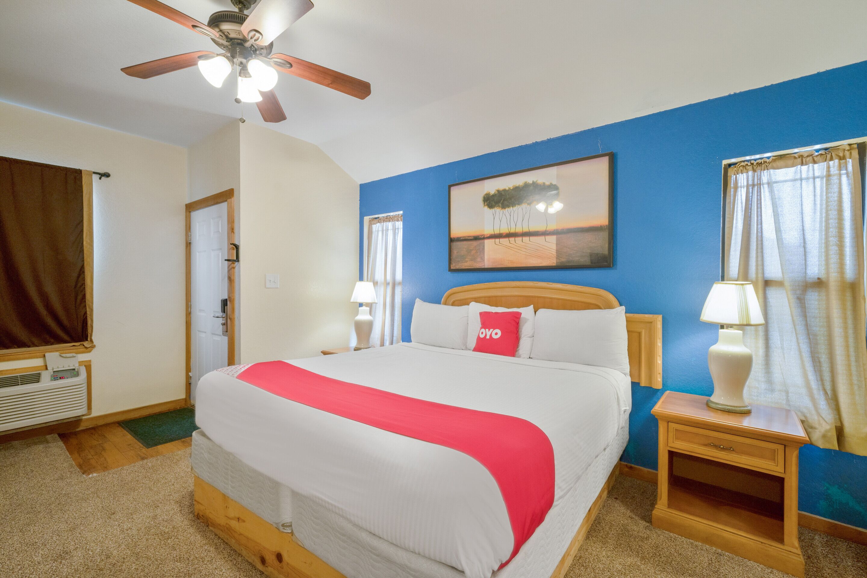 A hotel room with a bed, a ceiling fan, a nightstand, a lamp, a window with curtains, a door, and a painting on the wall.