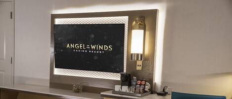 Una habitación de hotel moderna con una gran pantalla de televisión mostrando 'Angel of the Winds Casino Resort', un escritorio con cafetera y una silla azul.