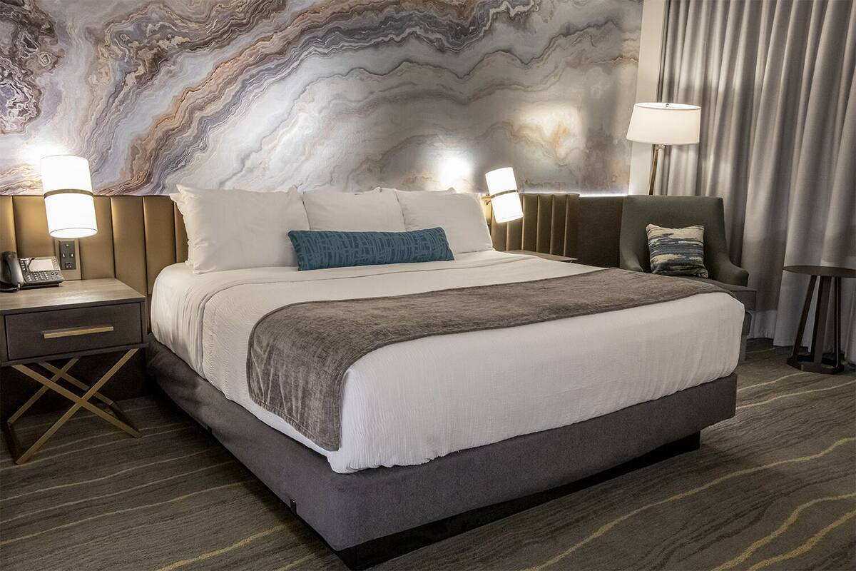 Een hotelkamer met een groot bed, twee nachtkastjes met lampen, een marmeren wand en een grijze gordijn.