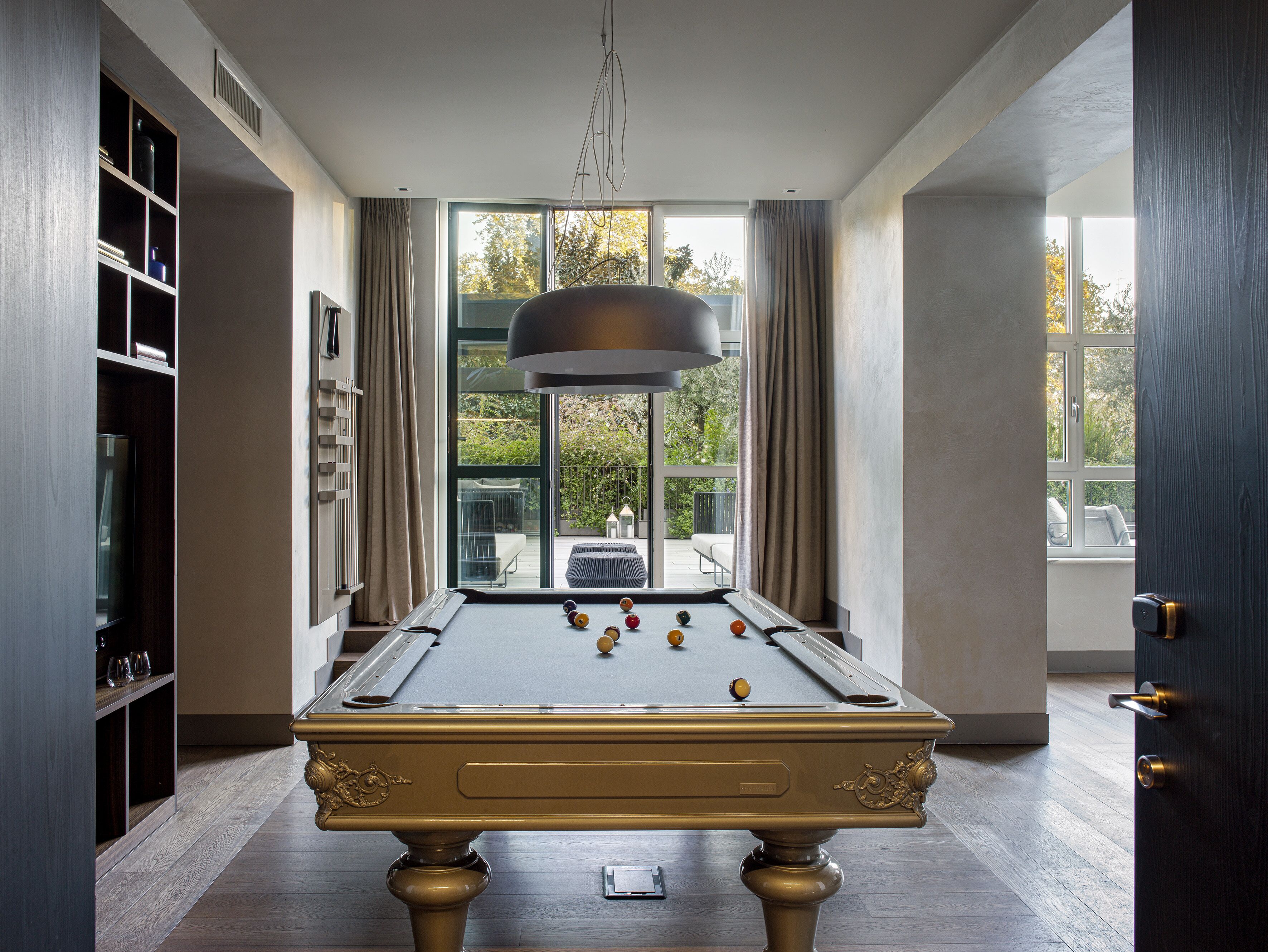 A pool table with billiard balls, a bookshelf, and a view through large windows.