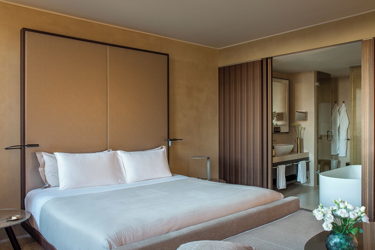 A hotel room with a large bed, a bedside table, a mirror, a bathroom with a shower, and a vase with flowers.