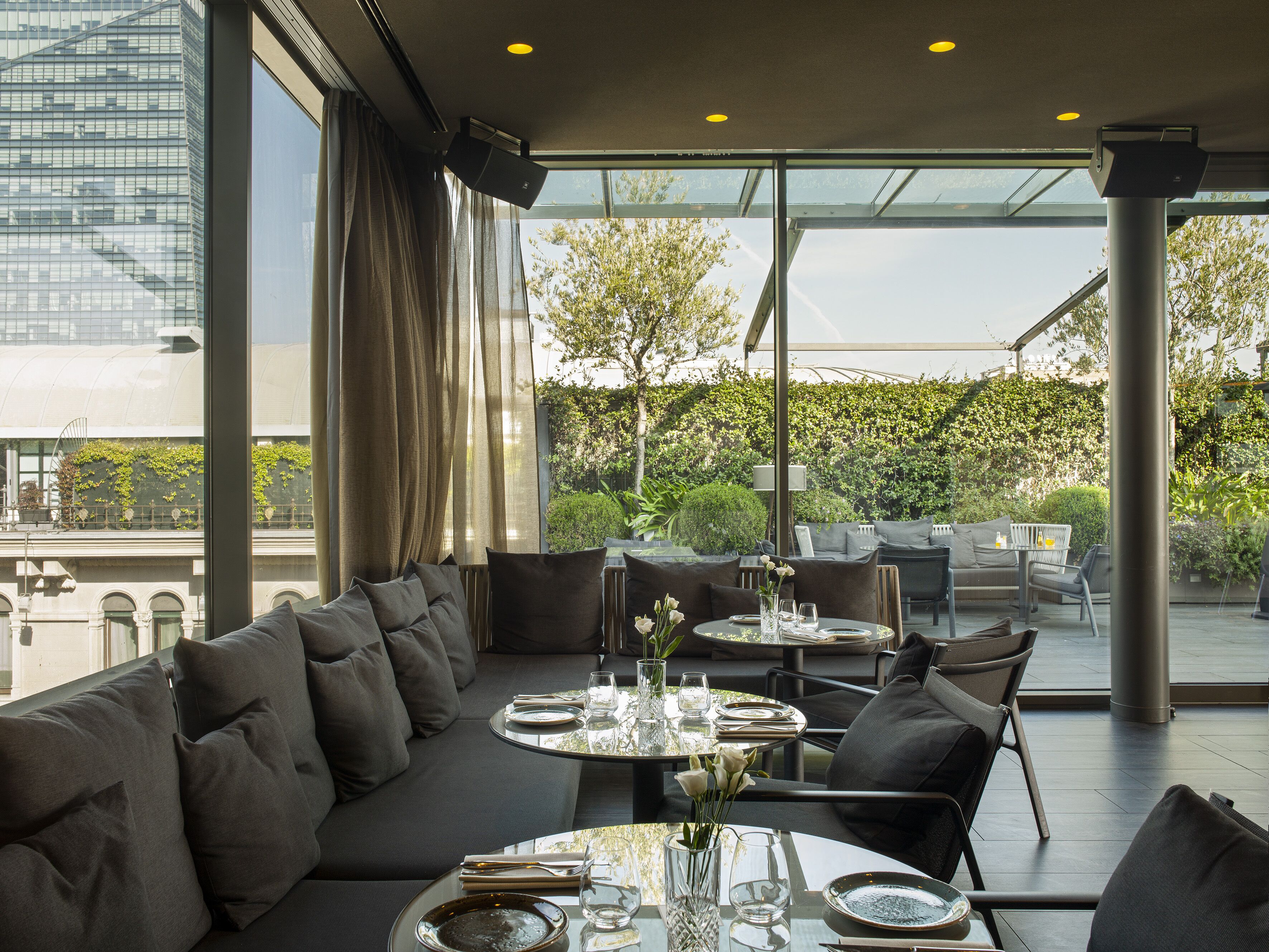 A modern outdoor dining area with a sofa, tables, and chairs, overlooking a cityscape and greenery.