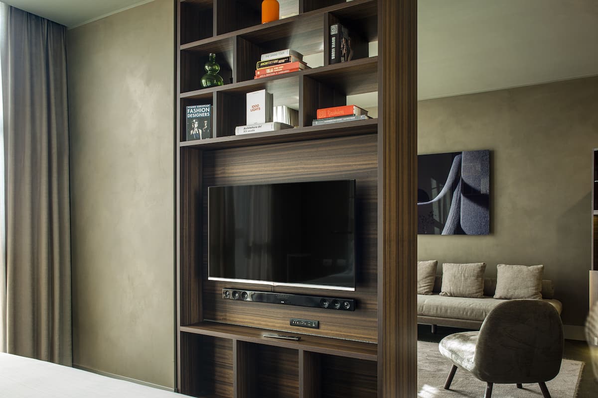 A modern hotel room with a large flat-screen TV, a wooden bookshelf with books and decorative items, a comfortable sofa, and a large window with curtains.