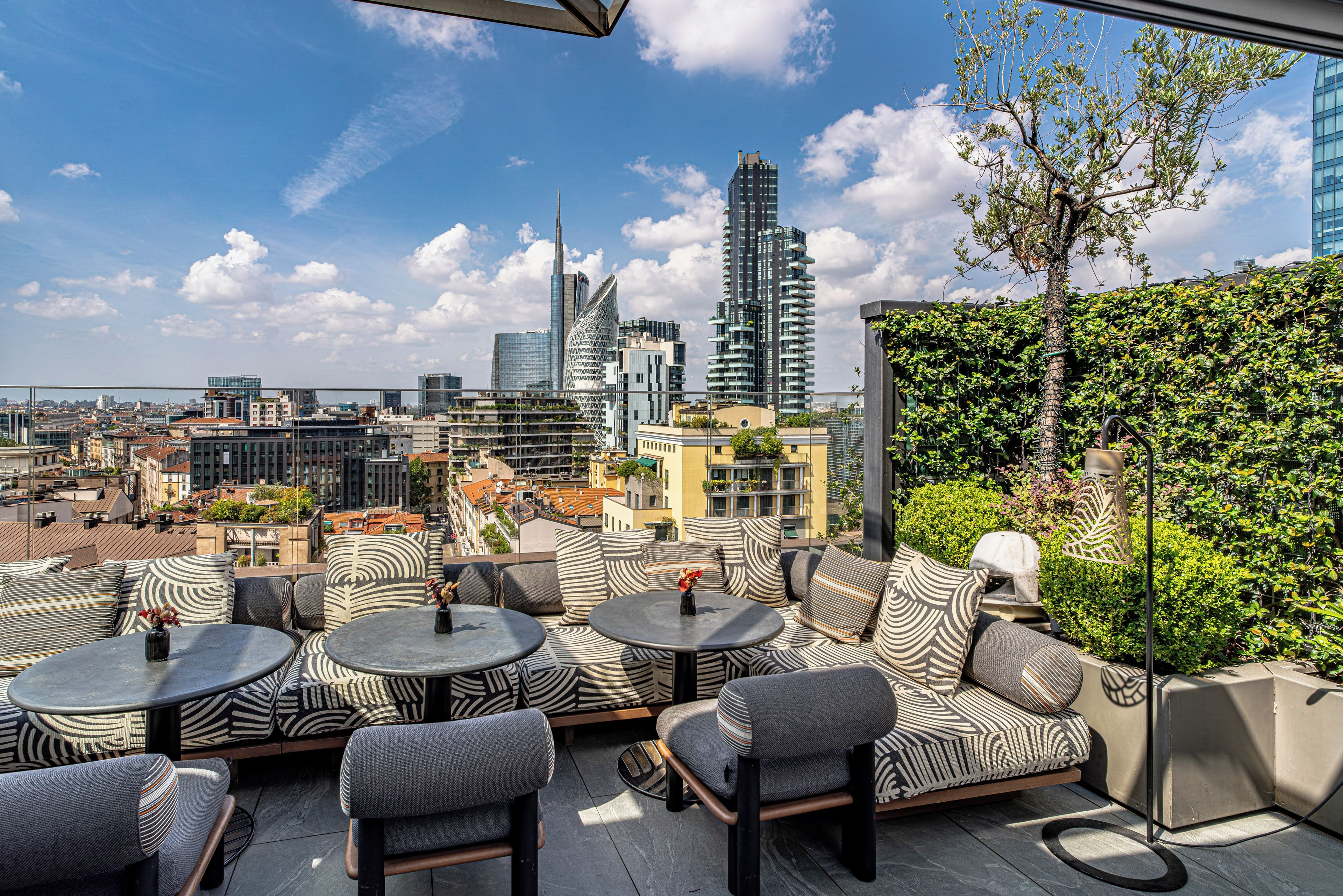 An outdoor seating area with round tables and cushioned chairs, overlooking a cityscape with modern buildings and a clear sky.