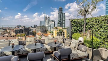 An outdoor seating area with round tables and cushioned chairs, overlooking a cityscape with modern buildings and a clear sky.