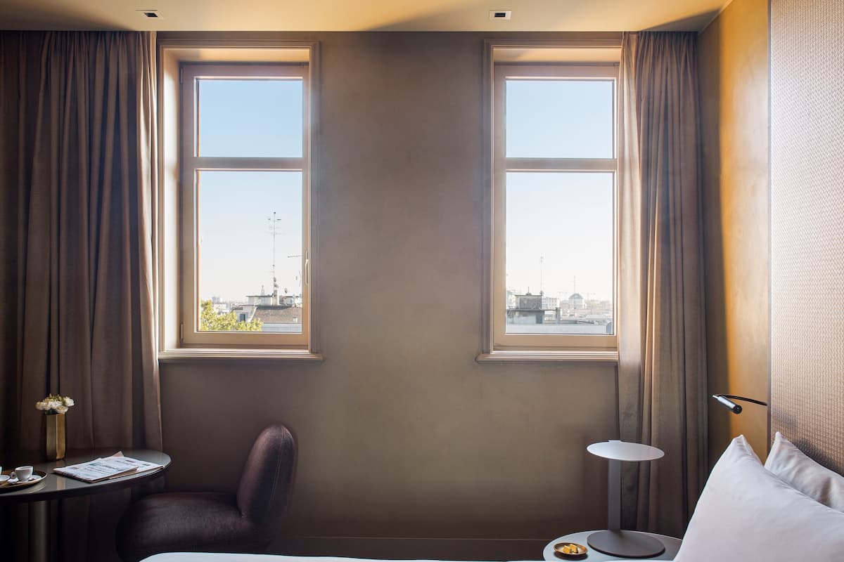 A hotel room with a bed, two windows, a chair, a small table with a lamp, and a view of the cityscape.