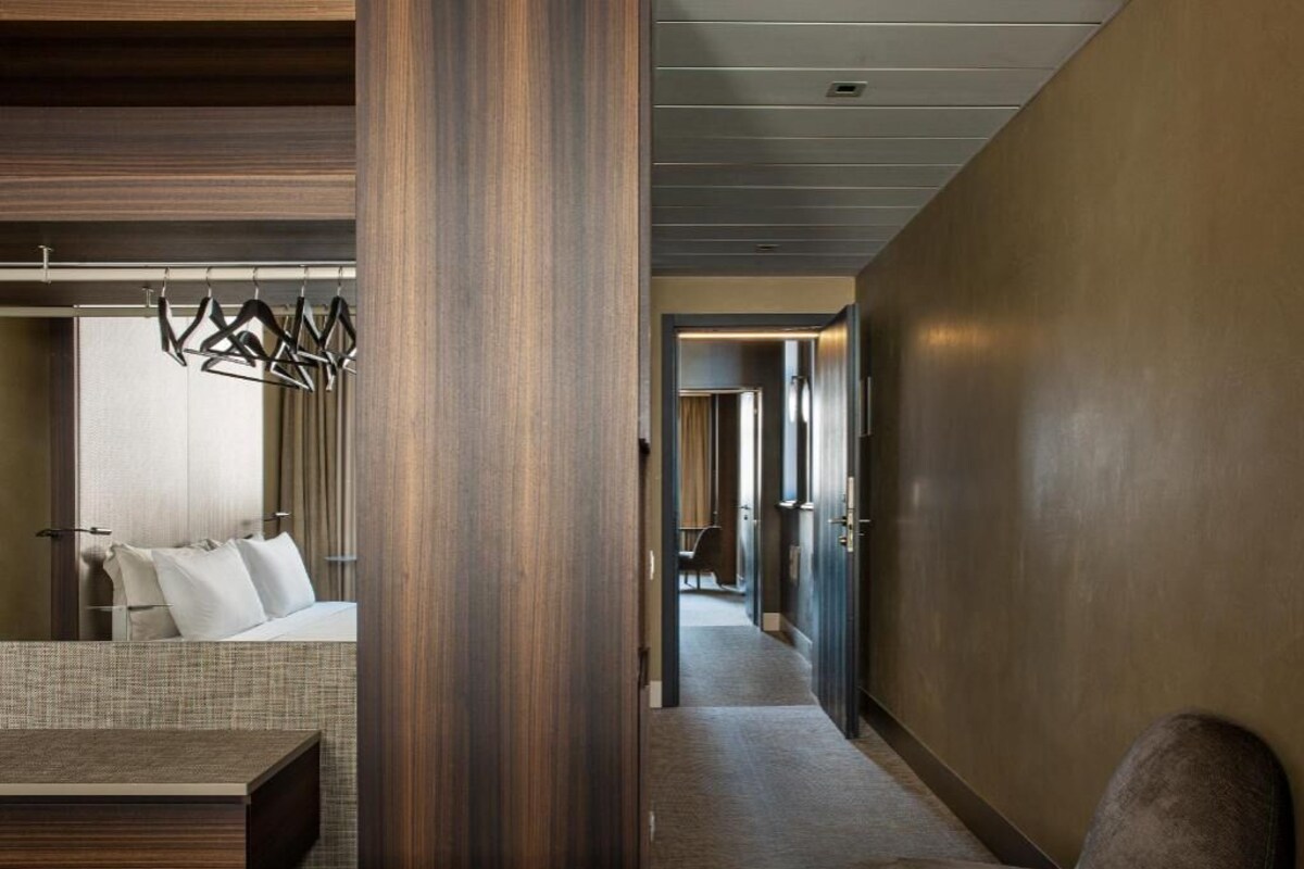 Habitación de hotel con una cama, cabecero de madera, zona de estar con un sofá y un pasillo que da a otras habitaciones.