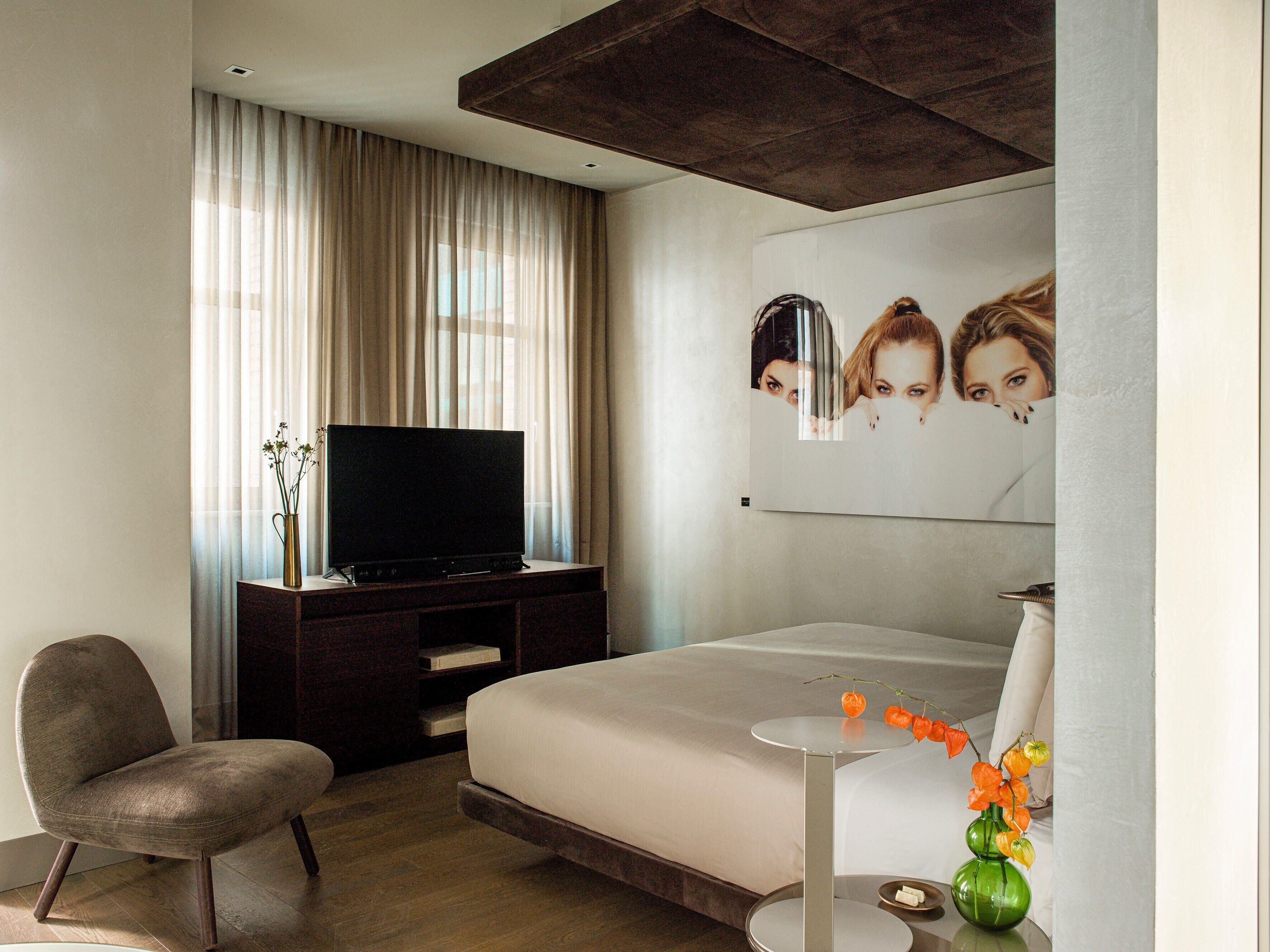 Una habitación de hotel moderna con una cama, un televisor sobre un soporte de madera, una silla y una mesita de noche con un jarrón de flores.