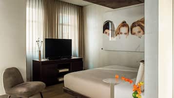 Una habitación de hotel moderna con una cama, un televisor sobre un soporte de madera, una silla y una mesita de noche con un jarrón de flores.