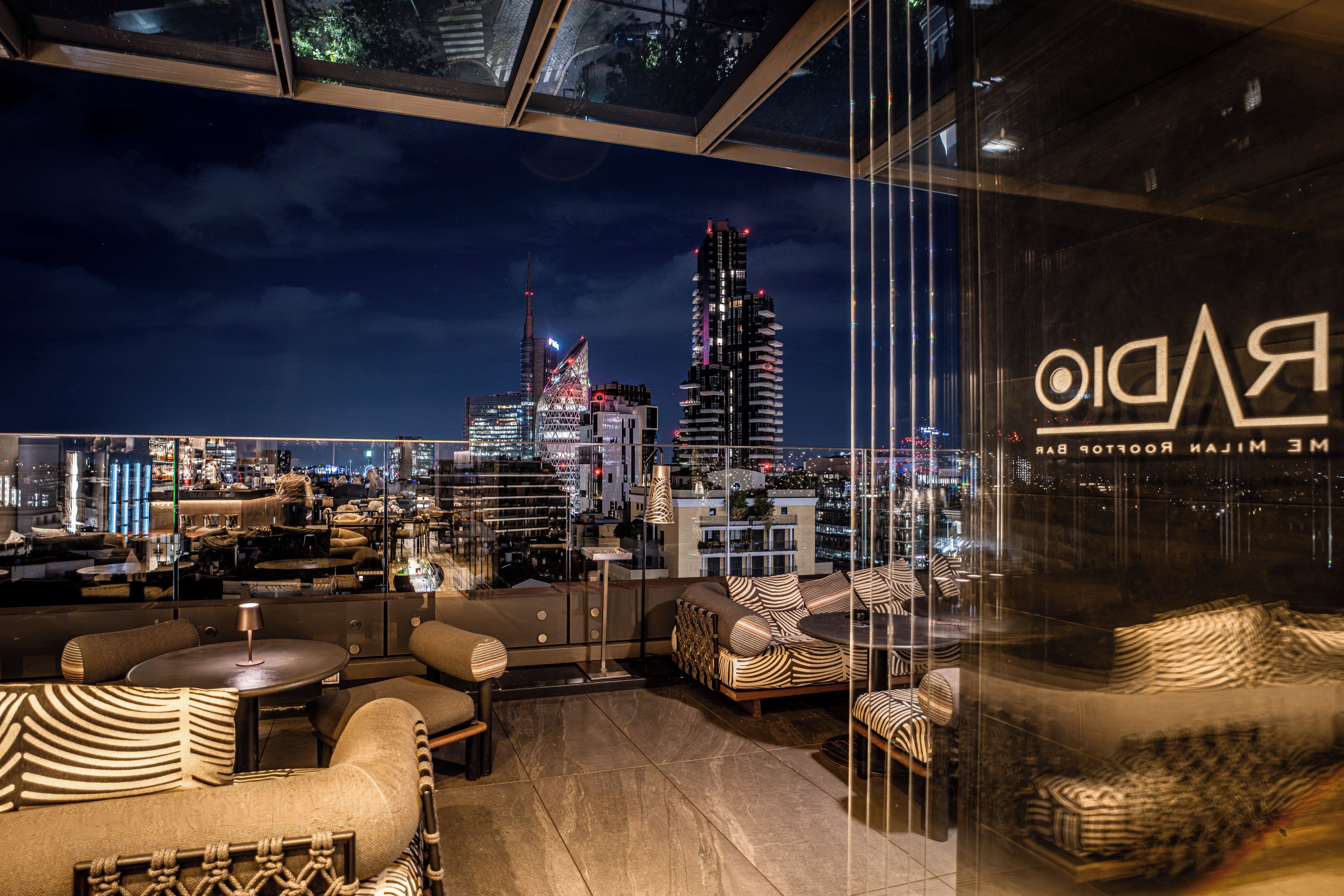 Un bar en la azotea con vista al horizonte urbano, amueblado con sofás y mesas, y una pared de vidrio que refleja las luces de la ciudad.