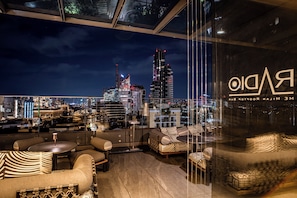 A rooftop bar with a city skyline view, furnished with sofas and tables, and a glass wall reflecting the city lights. - ME Milan - Il Duca (Milan)