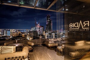 Un bar sul tetto con vista sullo skyline cittadino, arredato con divani e tavoli, e una parete vetrata che riflette le luci della città.