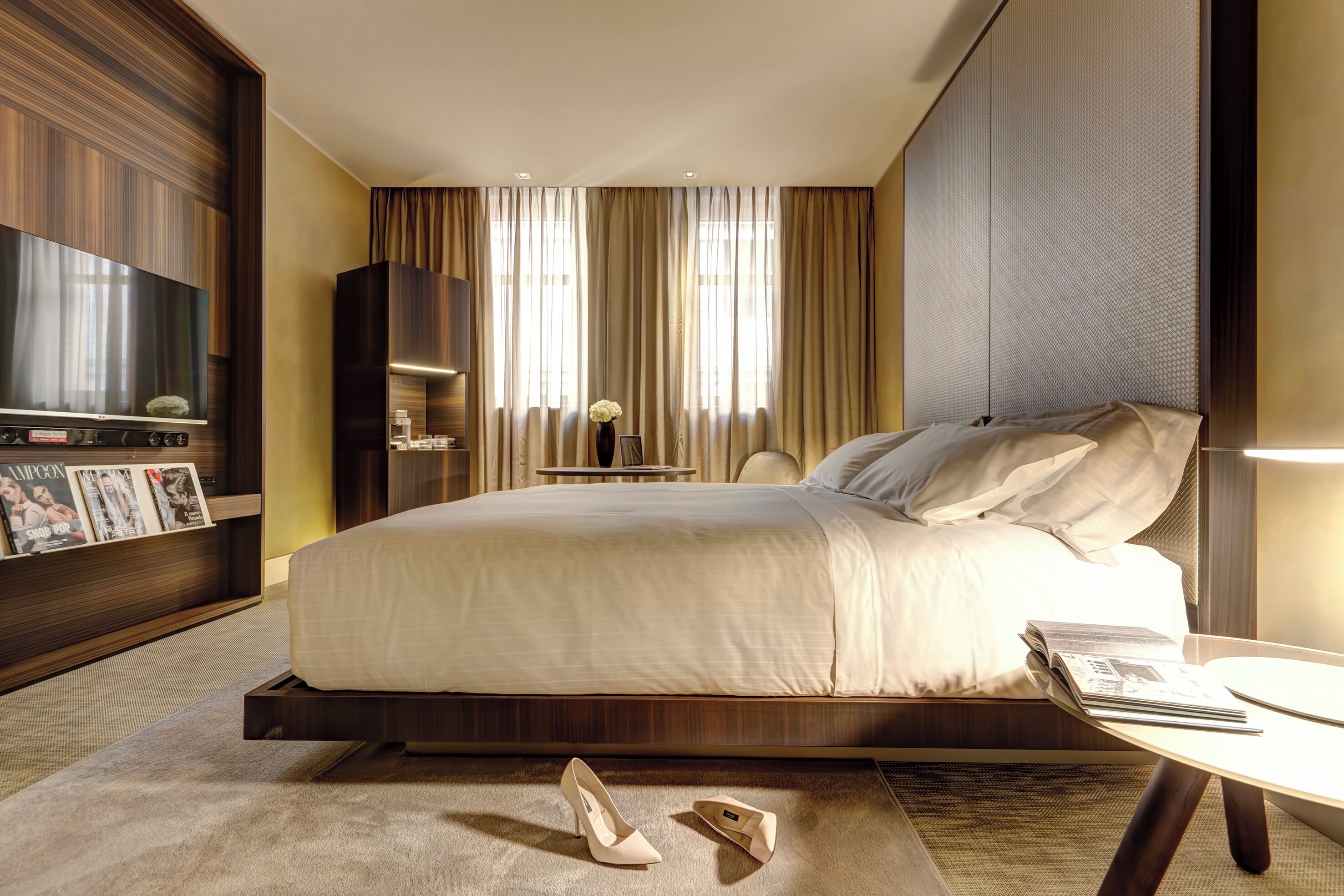 A modern hotel room with a large bed, a flat-screen TV, a bedside table, and a small side table with a vase.