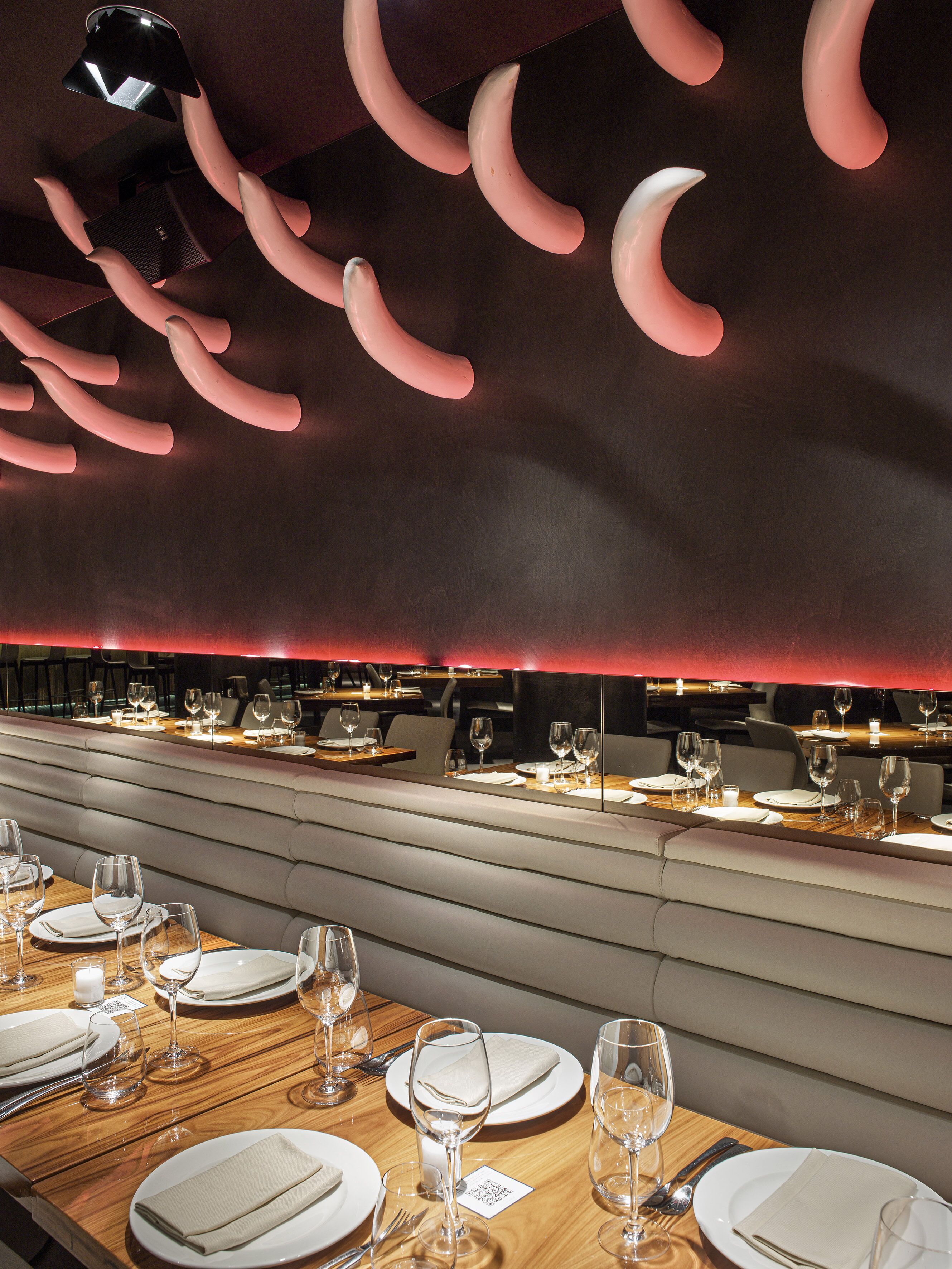 A restaurant with a modern interior, featuring a long dining table with multiple chairs, a high ceiling with unique lighting fixtures, and a red accent wall.