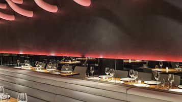 A restaurant with a modern interior, featuring a long dining table with multiple chairs, a high ceiling with unique lighting fixtures, and a red accent wall.