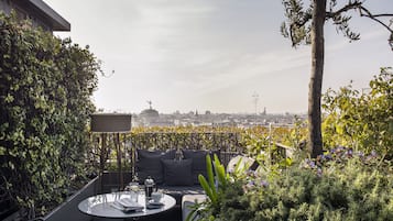 Terraza en la azotea con zona de estar, una mesa con periódicos y una cafetera, rodeada de vegetación y vistas a la ciudad.