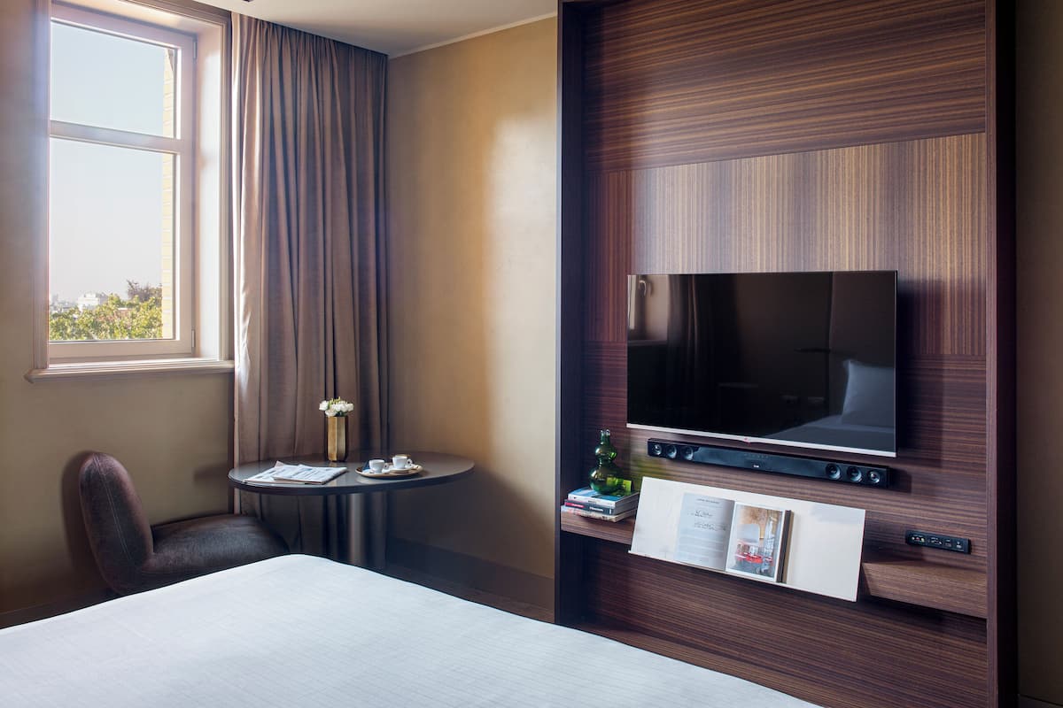 A hotel room with a bed, a television mounted on the wall, a small table with a cup and saucer, and a window with curtains.
