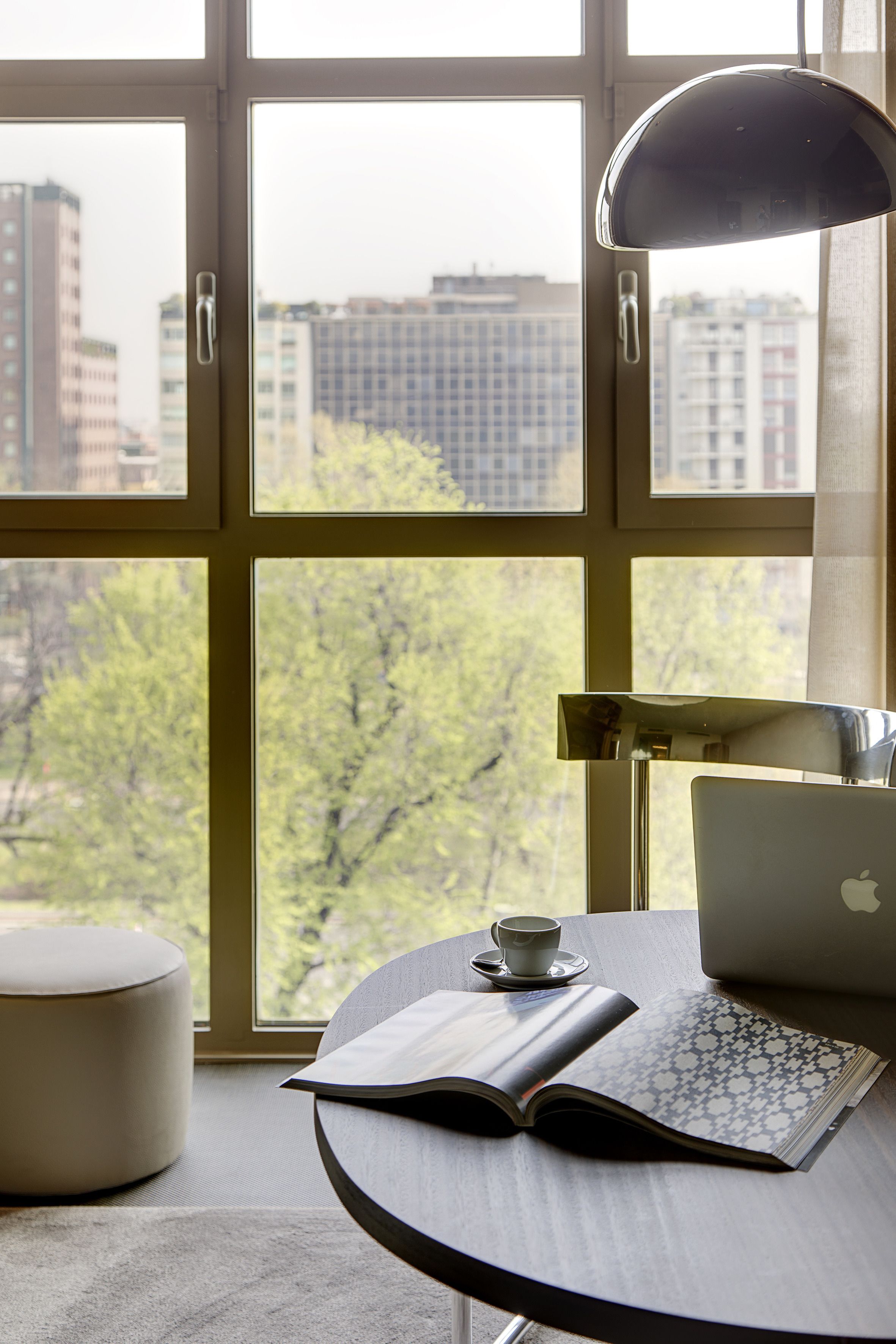 Una habitación con un gran ventanal, un escritorio con una computadora portátil, un libro y una taza.
