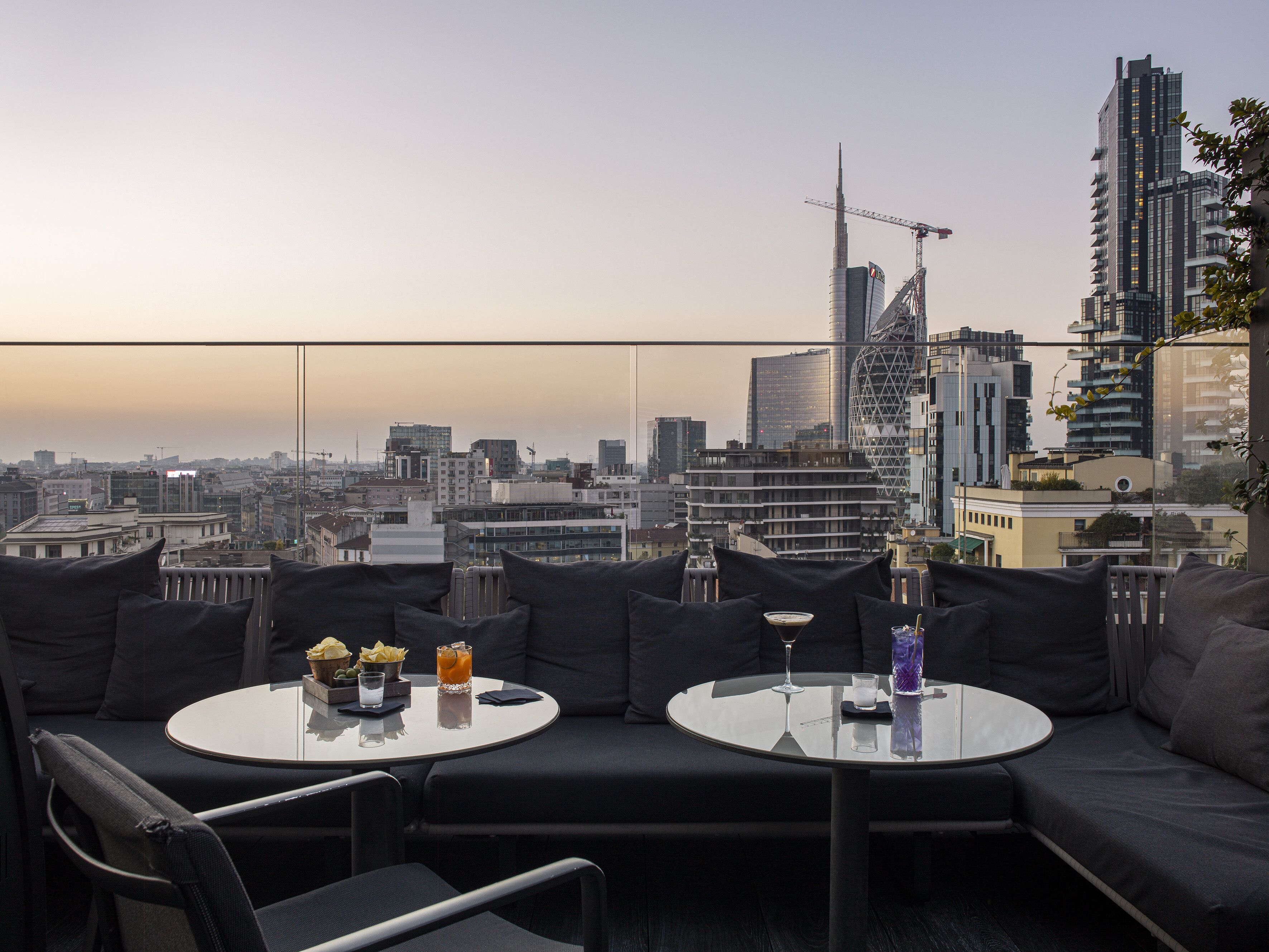 Eine Dachterrasse mit zwei runden Tischen, schwarzen Sitzgelegenheiten mit Polstern und Blick auf die Stadt.