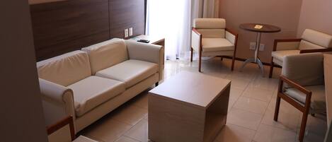 A modern hotel room with a beige sofa, two armchairs, a small round table, and a wooden headboard.