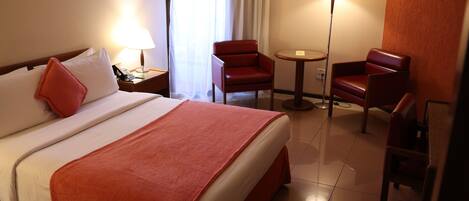 A hotel room with a bed, two chairs, a small table, a lamp, and a painting on the wall.