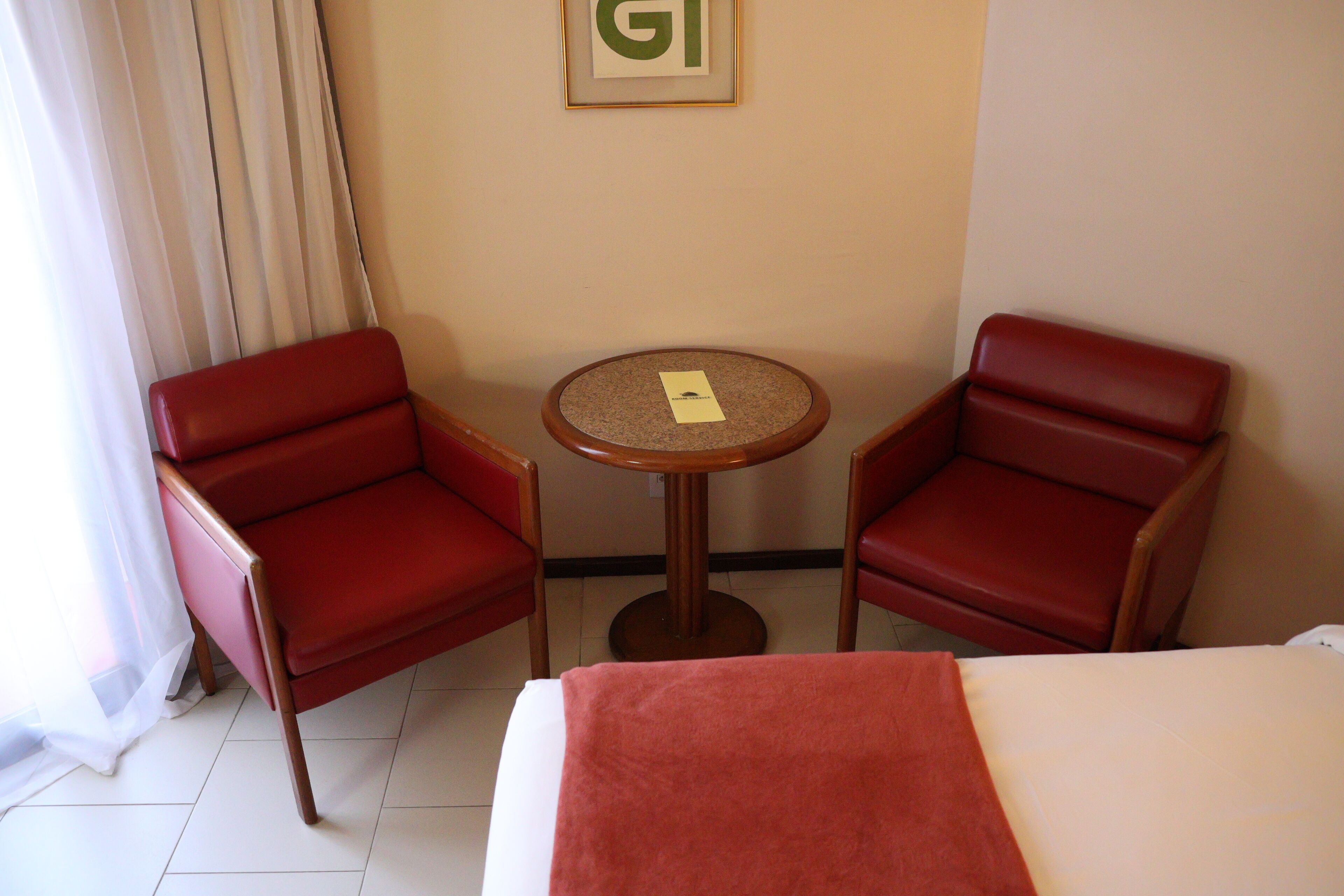 Une chambre d’hôtel avec deux fauteuils en cuir rouge, une petite table ronde et un lit recouvert d’une couverture blanche et rouge.