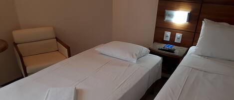 Habitación de hotel con dos camas, una silla, una mesita de noche con control remoto y una lámpara de pared.
