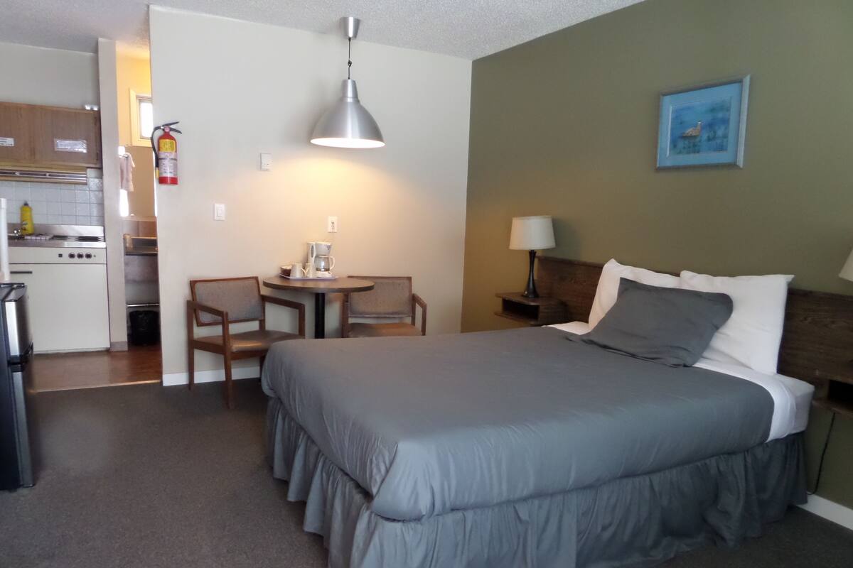 A compact hotel room with a bed, a small table with chairs, a kitchenette, and a painting on the wall.