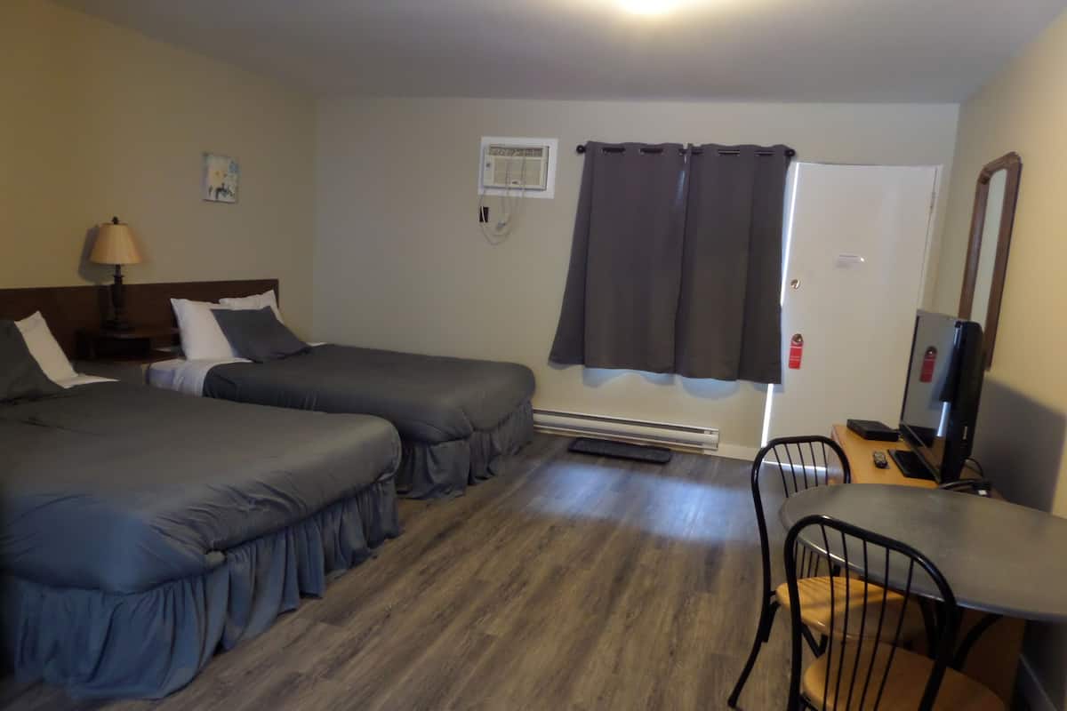 A hotel room with two beds, a desk with a chair, a lamp, a heater, a mirror, and a window with curtains.