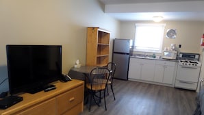 Una kitchenette compacta con refrigerador, microondas, estufa y pileta. Hay una zona de comedor con mesa y sillas, y un televisor sobre un mueble de madera.