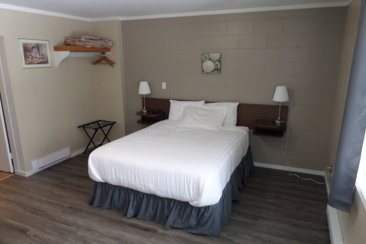 A neatly made bed with a white comforter and dark skirt, two bedside lamps, a small table with a lamp, a framed picture on the wall, and a brick-patterned wall.