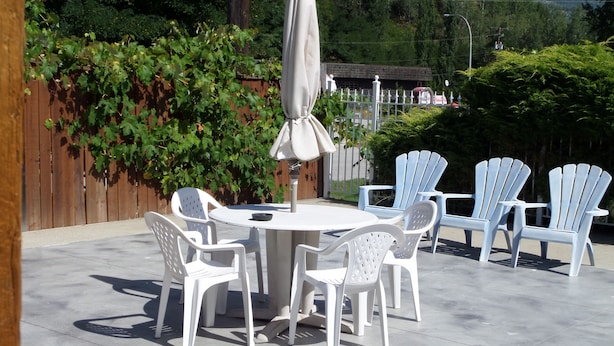 Un patio con una mesa redonda y seis sillas de plástico blancas, una sombrilla y una valla de madera con follaje verde.