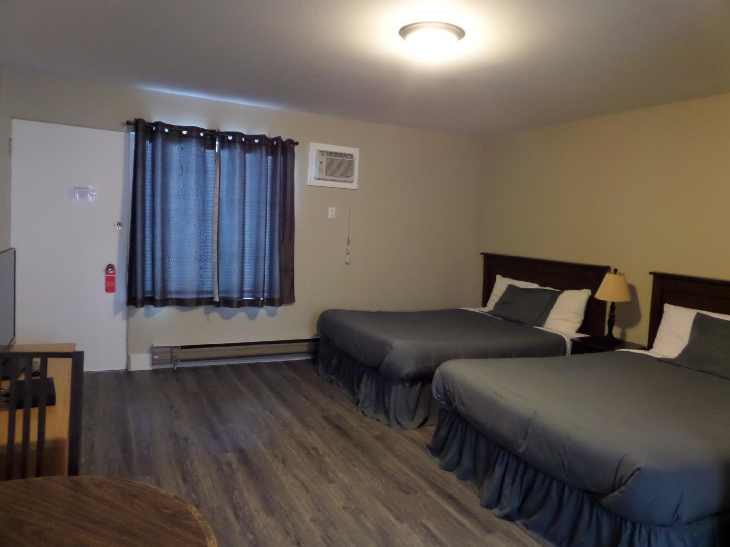 A hotel room with two beds, a wooden floor, a window with curtains, a heater, and a small table.