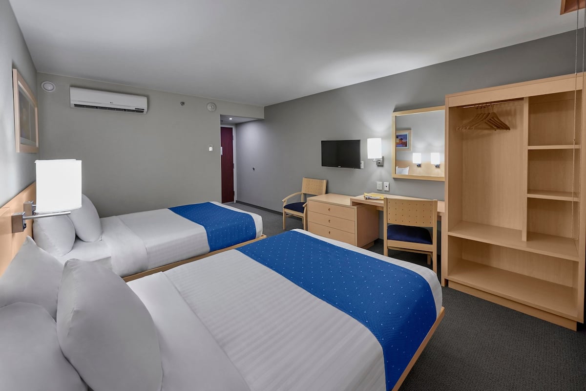 Habitación de hotel con dos camas, un escritorio, una silla, un armario, un televisor y aire acondicionado.